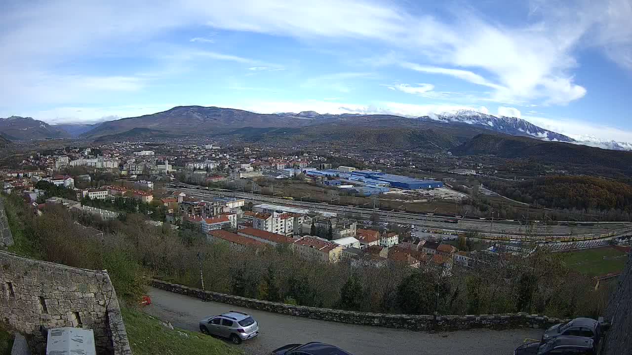 A wide panoramic view from a hillside reveals a town with a sprawling railway yard and various buildings, all nestled against a backdrop of rolling hills and distant snow-capped mountains under a bright, partly cloudy sky.