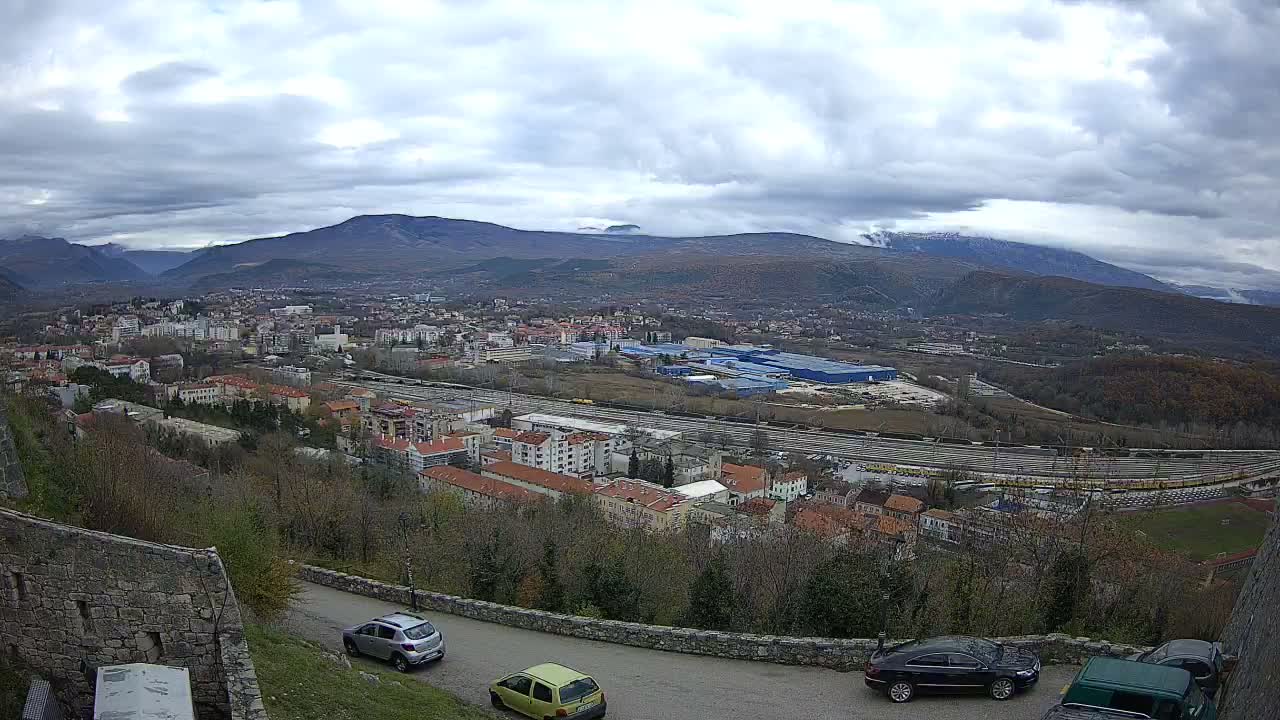An elevated panoramic shot captures a town spread across a valley, featuring residential buildings, a large railway yard, and industrial complexes, all set against a backdrop of mountains under a heavily overcast and gloomy sky with traces of snow on some distant peaks.