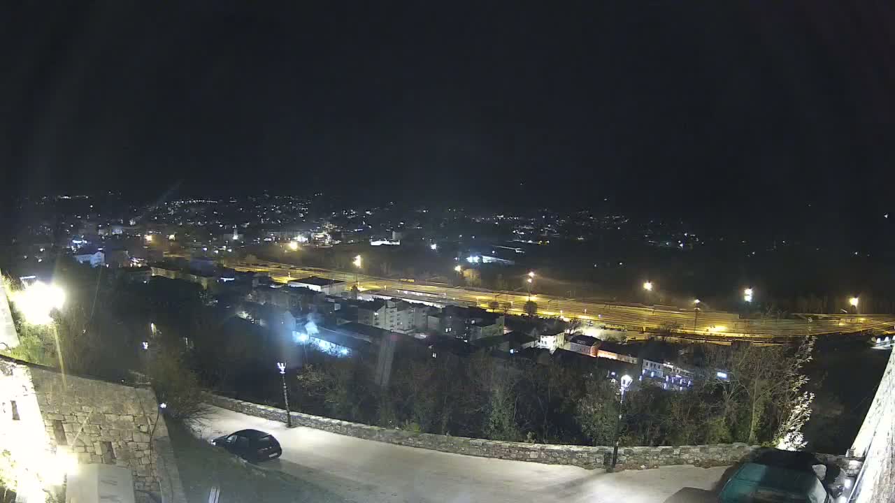 An elevated night view of a sprawling town under a clear sky shows a winding street with two parked cars in the foreground, overlooking residential buildings, a brightly lit highway, and distant city lights.