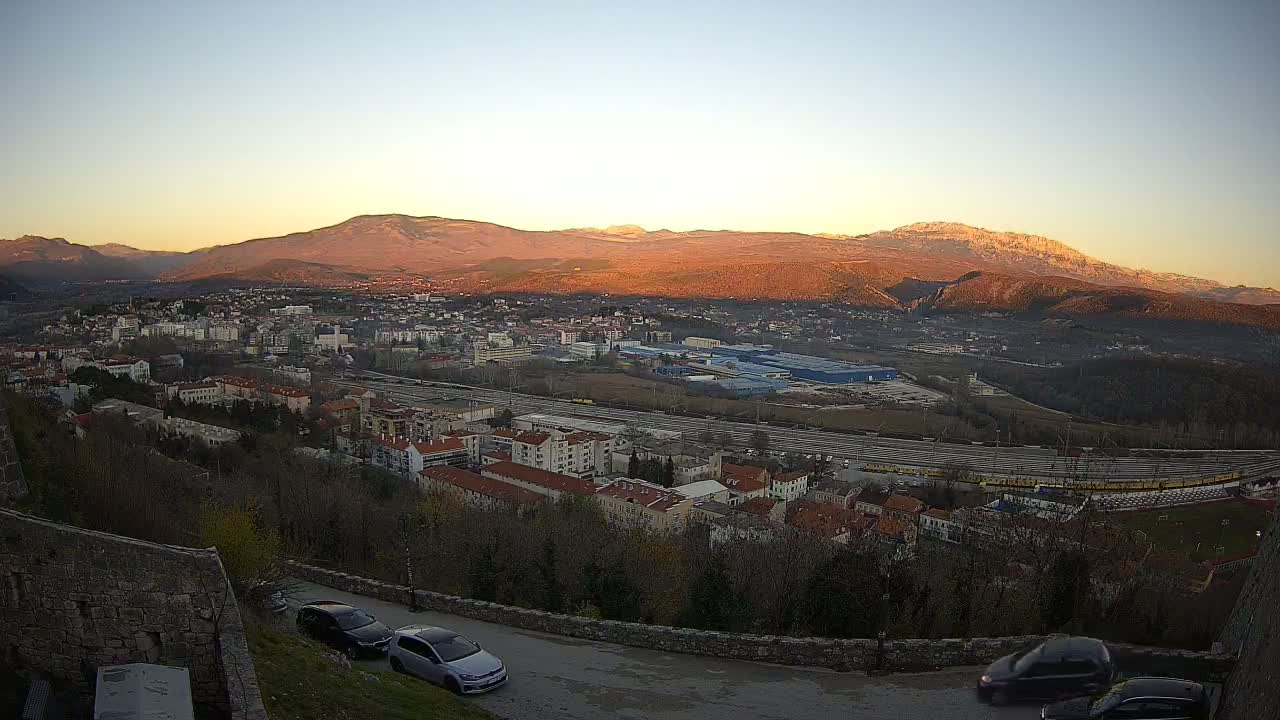 An elevated night view of a sprawling town under a clear sky shows a winding street with two parked cars in the foreground, overlooking residential buildings, a brightly lit highway, and distant city lights.