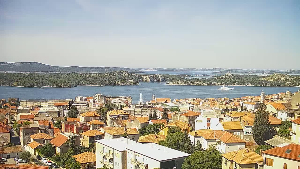 A sunny day reveals a coastal town with red-tiled roofs, nestled beside a calm blue bay dotted with islands under a clear sky.