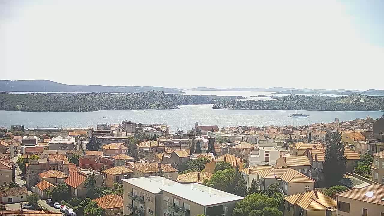 A coastal town with many low-rise buildings with orange roofs overlooks a calm bay and distant islands under a bright, mostly clear sky.