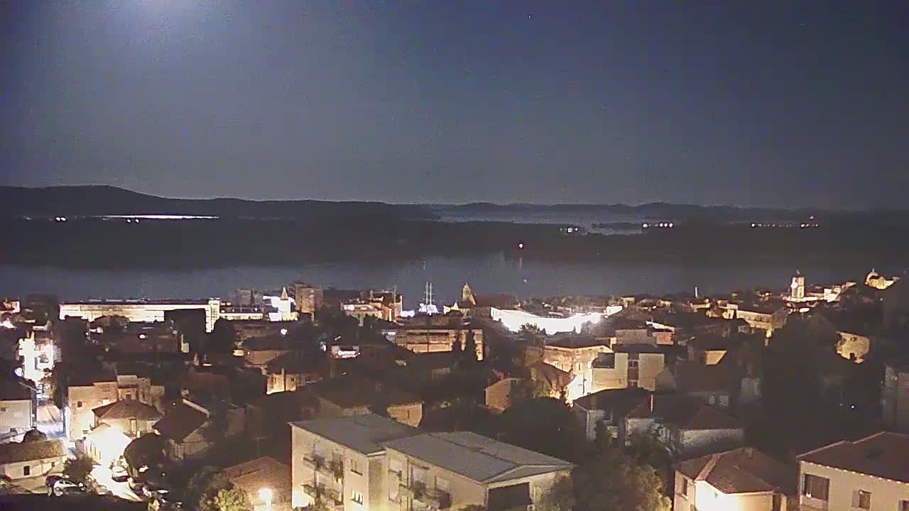 A nighttime view of a town illuminated by lights, overlooking a body of water partially obscured by fog under a bright moon.