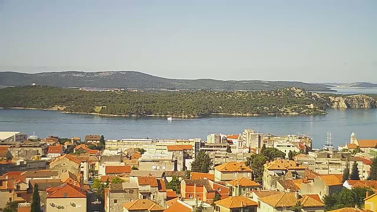 A coastal town with red-tiled roofs overlooks a calm body of water and a verdant island under a clear sky.