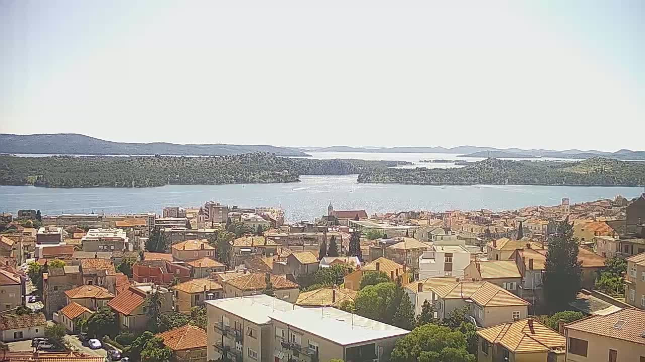 A coastal town with orange-tiled roofs overlooks a calm bay under a bright, sunny sky.