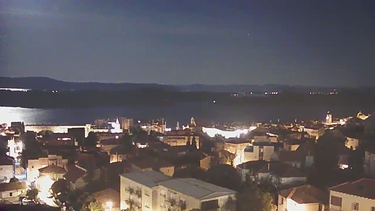 A nighttime view of a coastal town illuminated by lights, overlooking a calm body of water under a clear, dark sky.