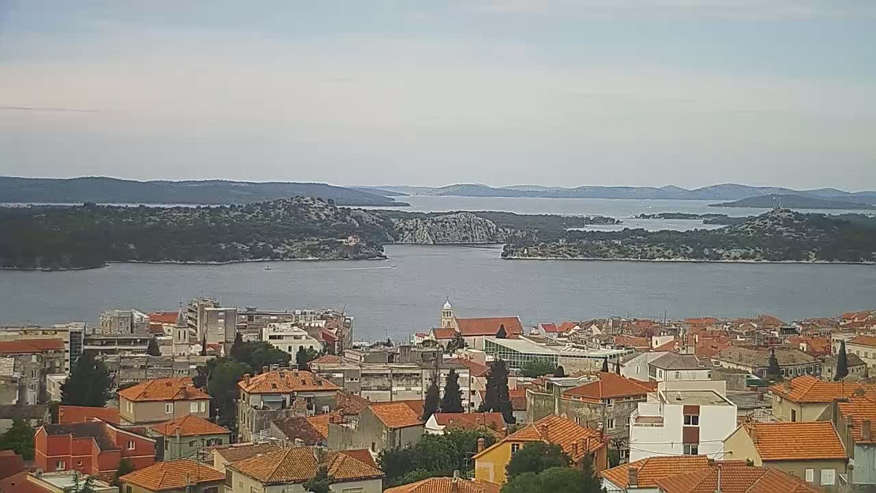 A coastal town with red-tiled roofs overlooks a calm body of water dotted with islands under a cloudy sky.