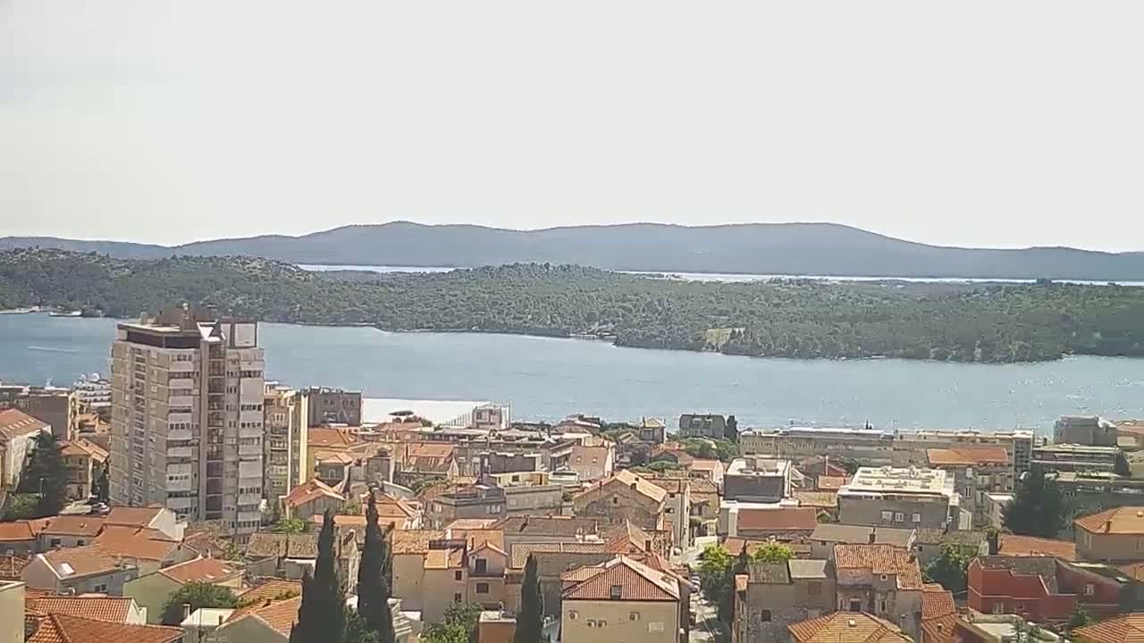 A coastal town with red-tiled roofs and a tall building overlooks a calm body of water and a distant, forested island under a bright, clear sky.