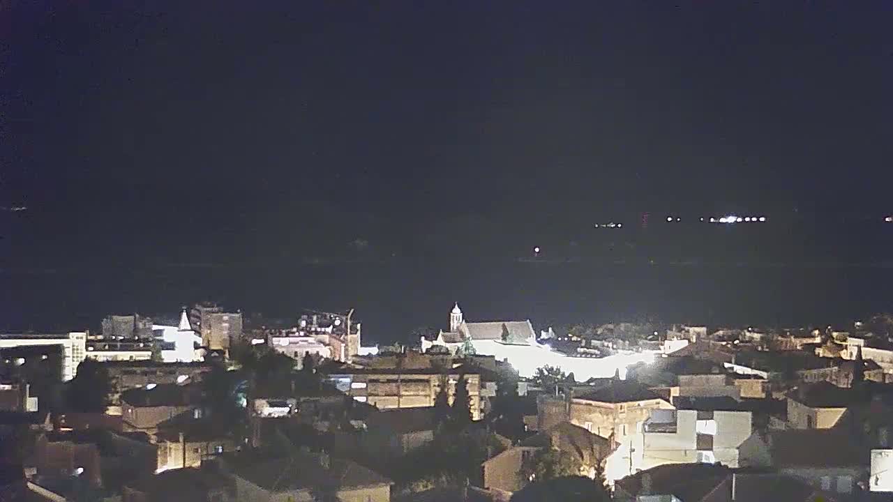 A nighttime, high-angle view shows a town's cityscape with a church and a body of water in the distance under a dark, clear sky.