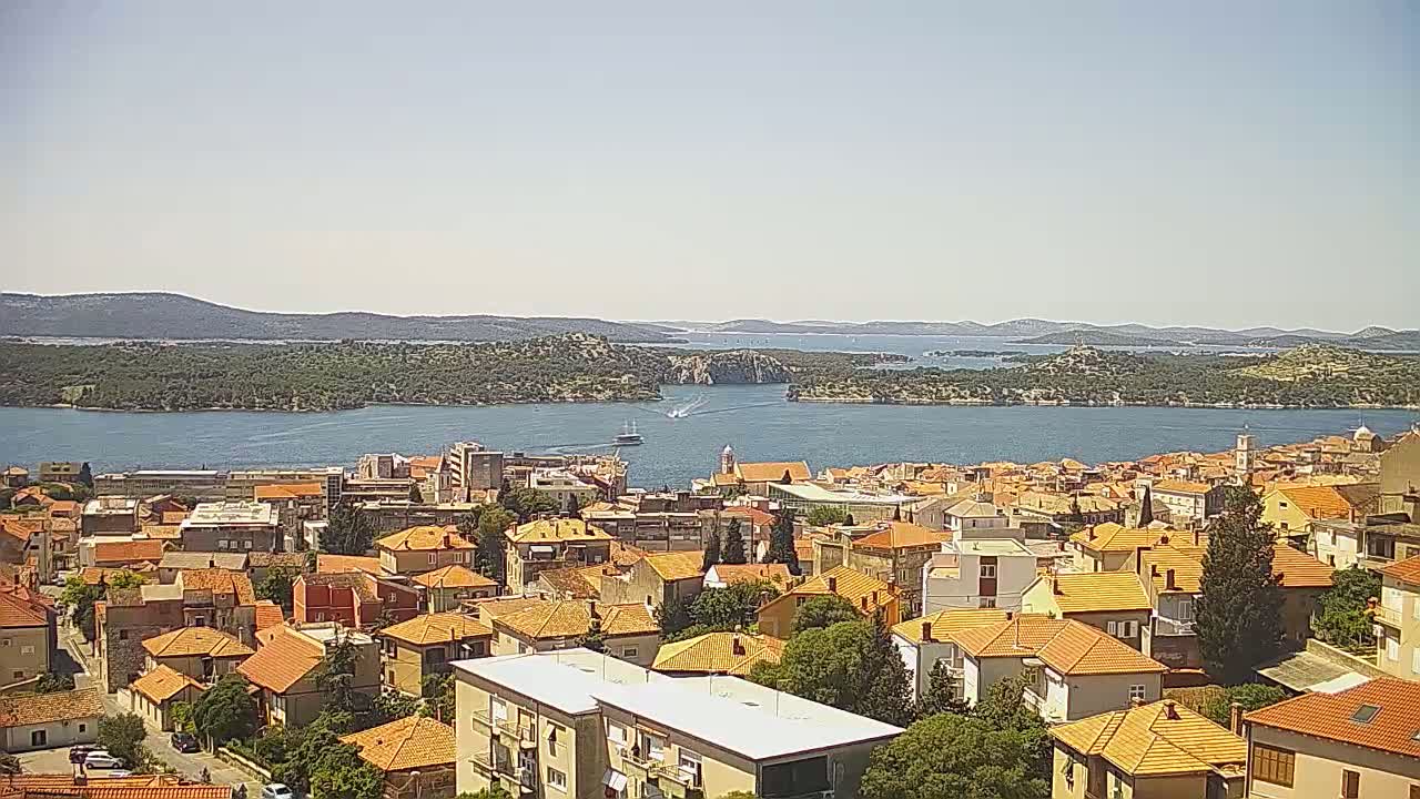 A coastal town with orange-tiled roofs overlooks a calm body of water on a sunny day, with islands and mountains visible in the distance.