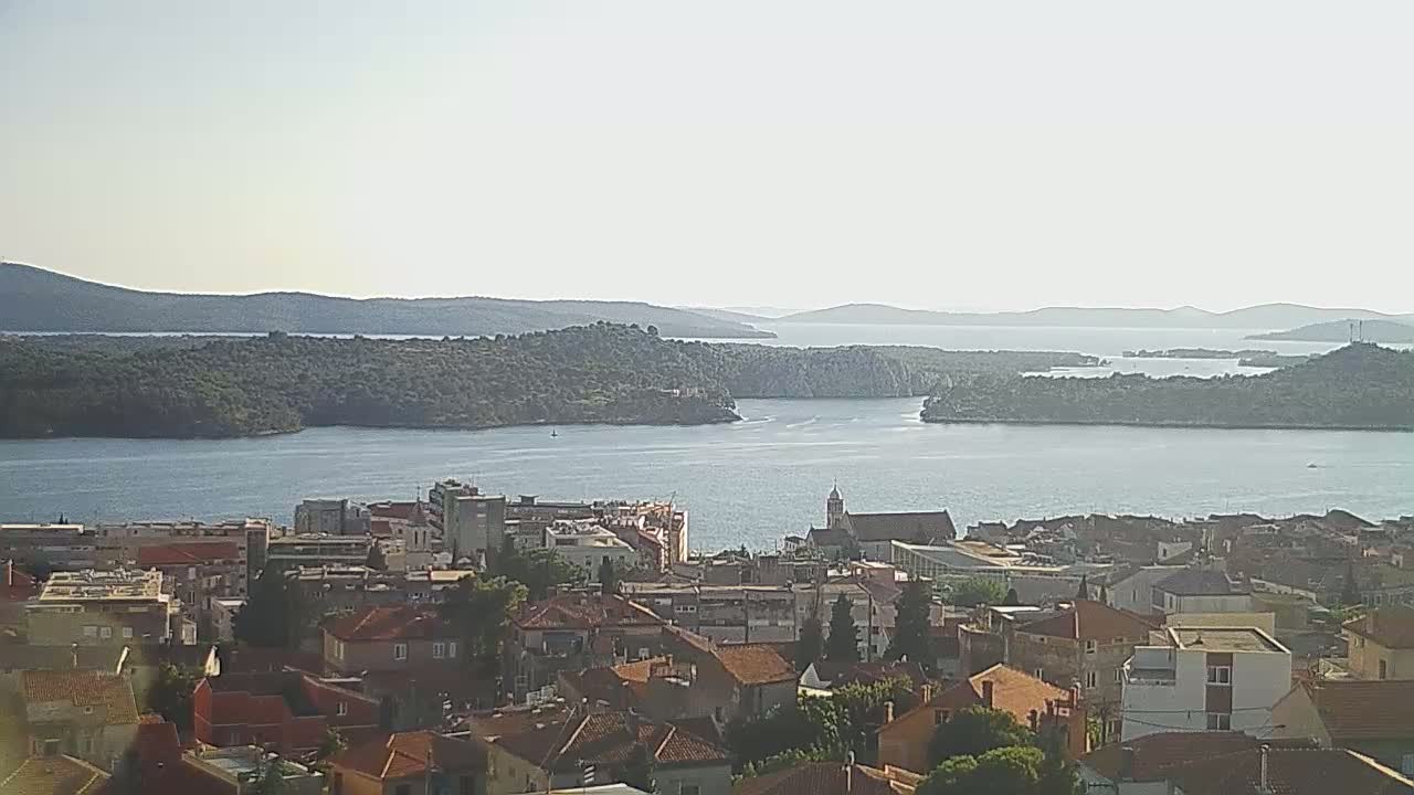 A coastal town with red-tiled roofs overlooks a calm bay surrounded by islands and mountains under a hazy sky.