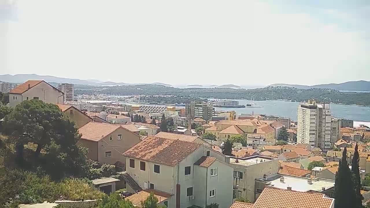 A coastal town with red-tiled roofs and a few taller buildings is visible on a sunny day, overlooking a calm body of water and distant islands.