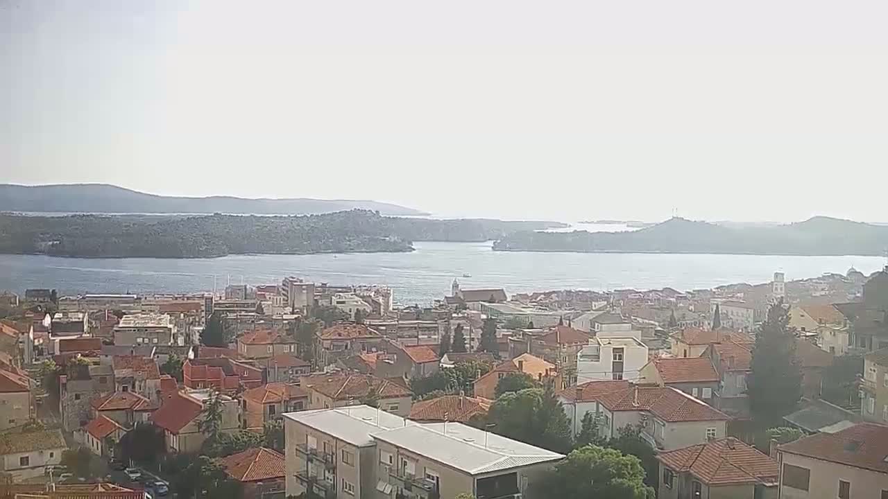 A hazy, sunny day reveals a coastal town nestled beside a calm bay, with distant islands and hills visible in the background.
