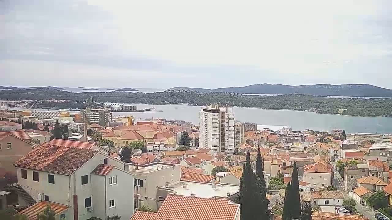 A coastal town with red-tiled roofs and a few taller buildings overlooks a calm bay under a mostly cloudy sky.
