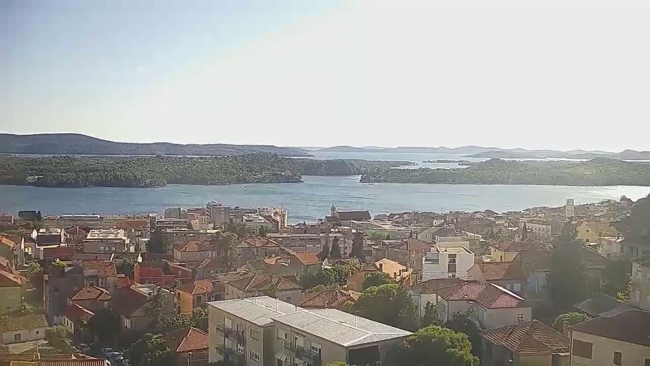 A coastal town with red-tiled roofs sprawls across a hillside overlooking a calm, sunlit bay dotted with islands under a clear sky.