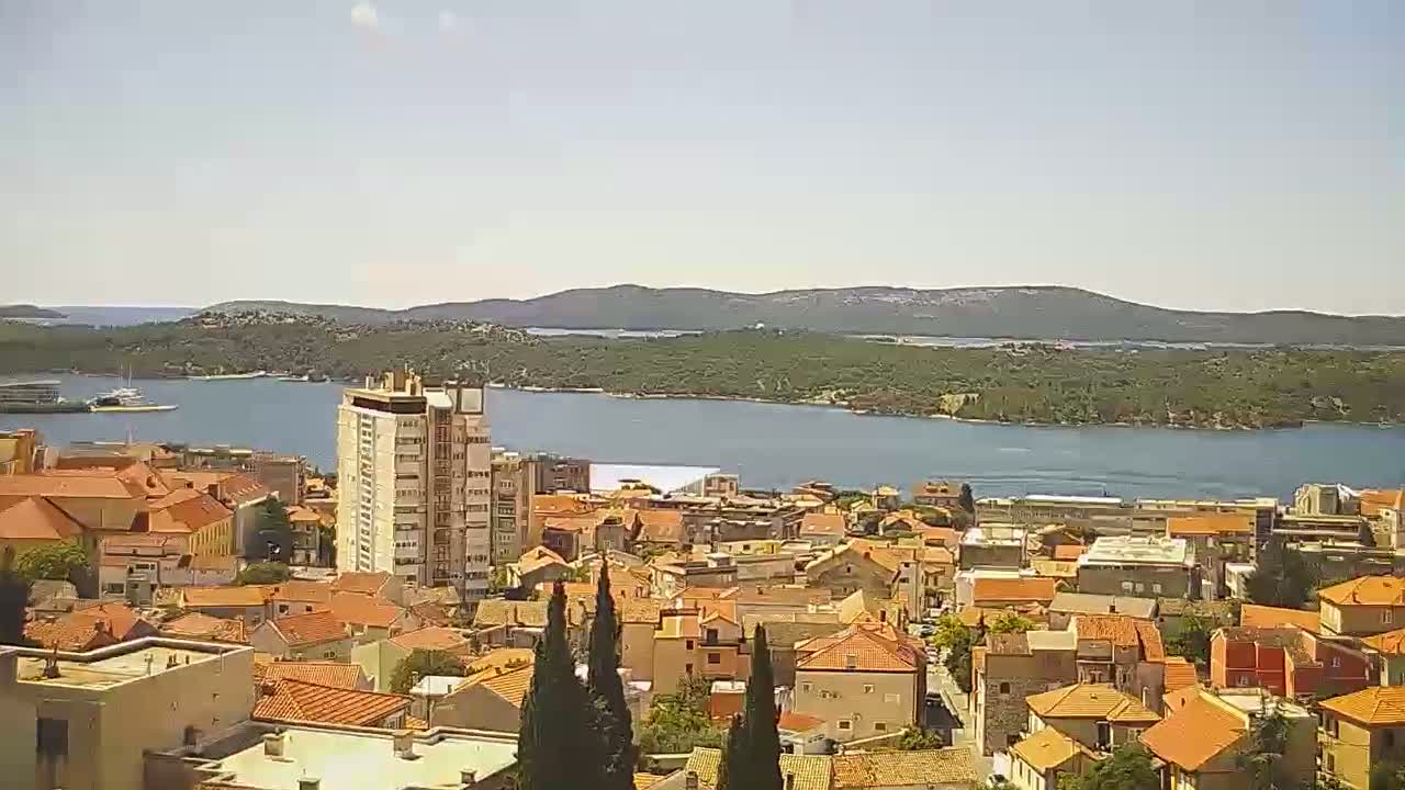 A coastal town with red-tiled roofs and a high-rise building overlooks a calm body of water under a mostly clear sky.