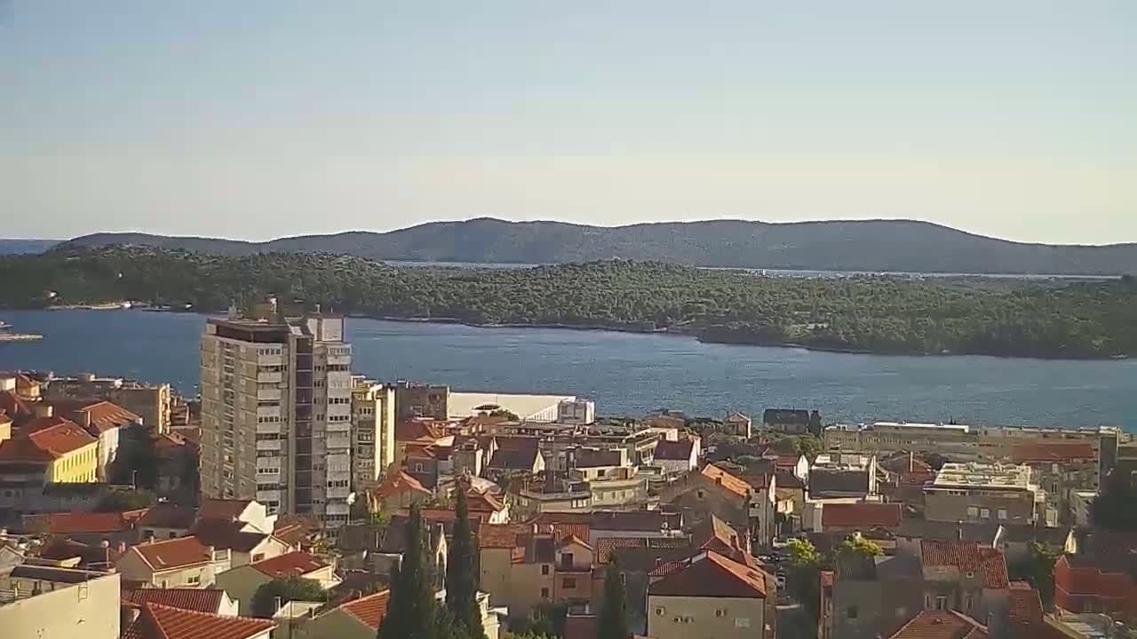A coastal town with red-tiled roofs and a tall building overlooks a calm body of water and a verdant island under a clear sky.