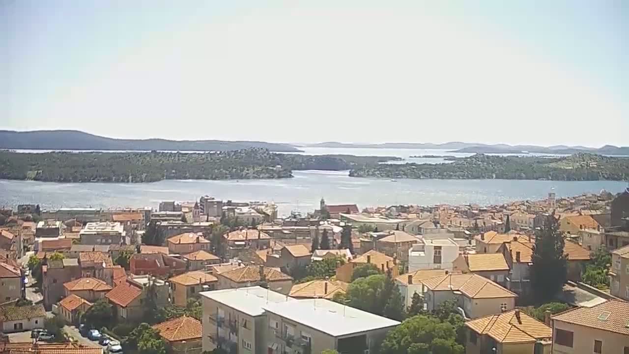 A coastal town with many orange-roofed buildings overlooks a calm body of water dotted with islands under a bright, sunny sky.