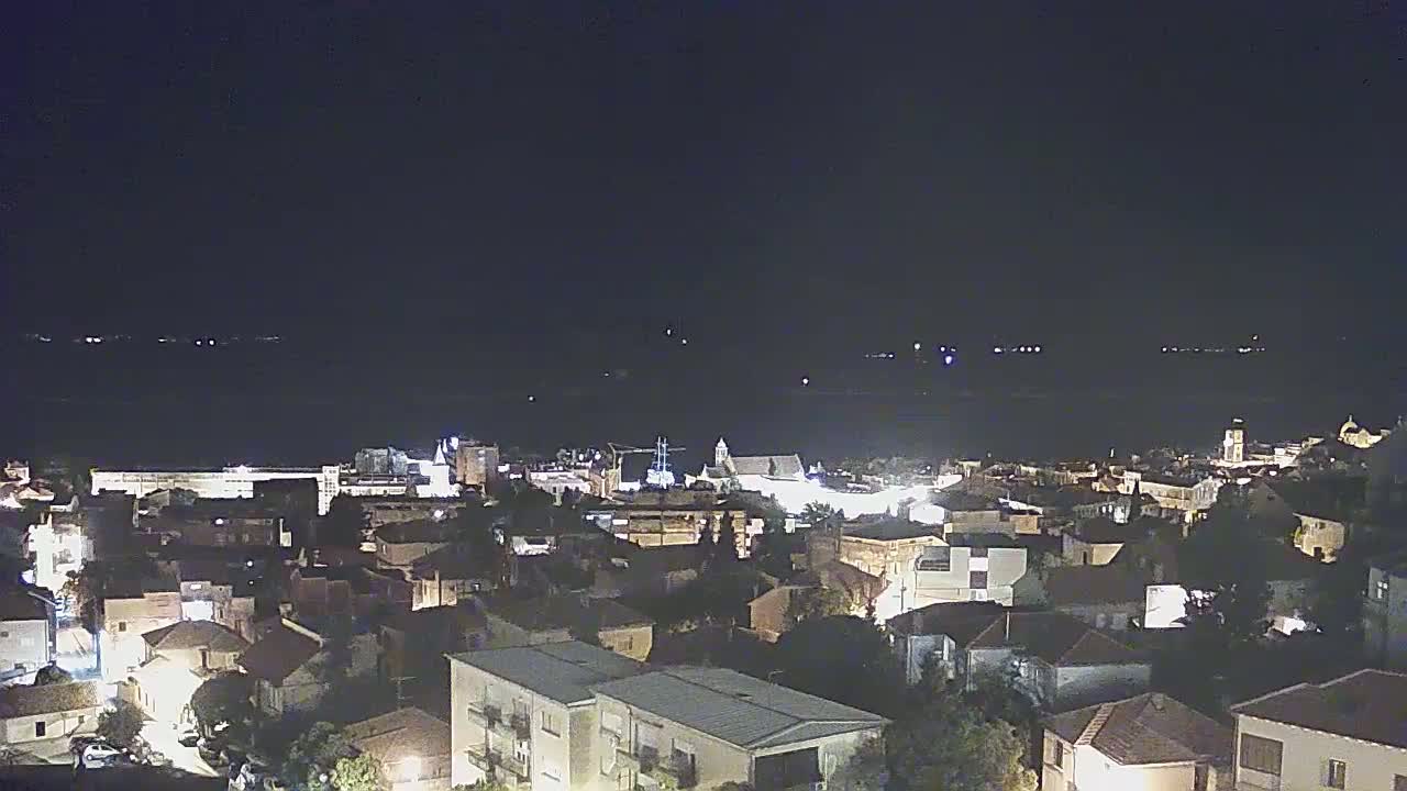 A nighttime view of a coastal town illuminated with lights, overlooking a dark body of water under a clear sky.