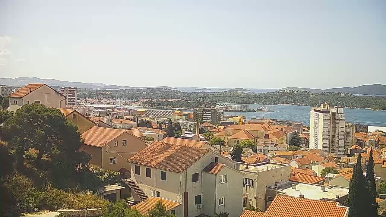 A coastal town with red-tiled roofs and a few taller buildings is visible on a sunny day, overlooking a calm body of water and distant islands.
