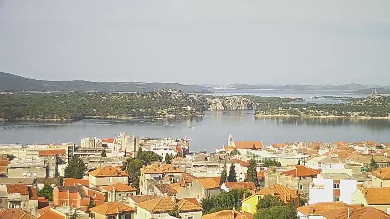 A coastal town with red-tiled roofs overlooks a calm body of water surrounded by green hills under a hazy sky.