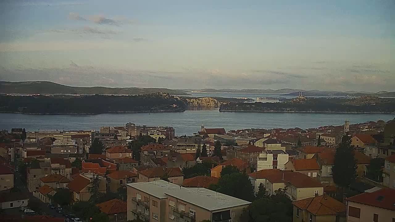 A coastal town with red-tiled roofs overlooks a calm bay and distant islands under a partly cloudy sky.