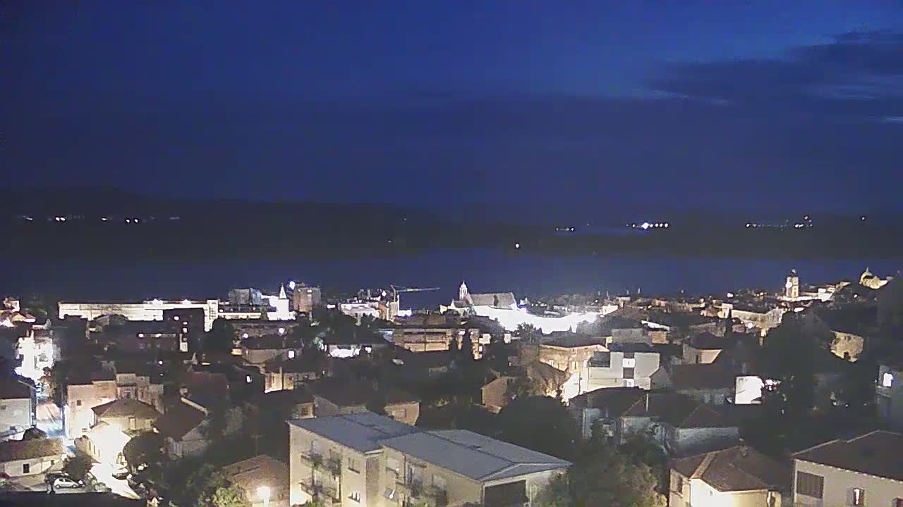 A nighttime view of a coastal town illuminated with streetlights, overlooking a dark body of water under a clear, dark blue sky.