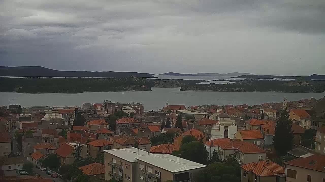 A coastal town with red-tiled roofs overlooks a calm body of water under a cloudy sky.