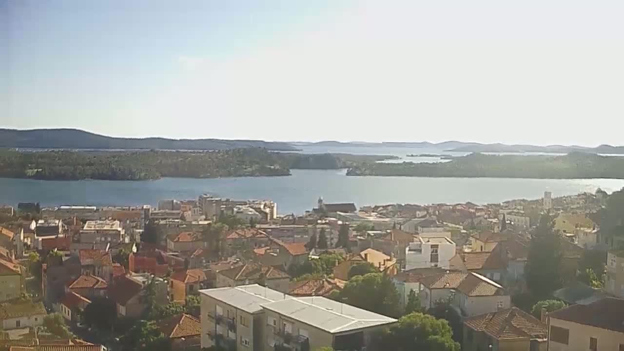 A coastal town with red-tiled roofs overlooks a calm bay surrounded by hills and islands under a bright, sunny sky.