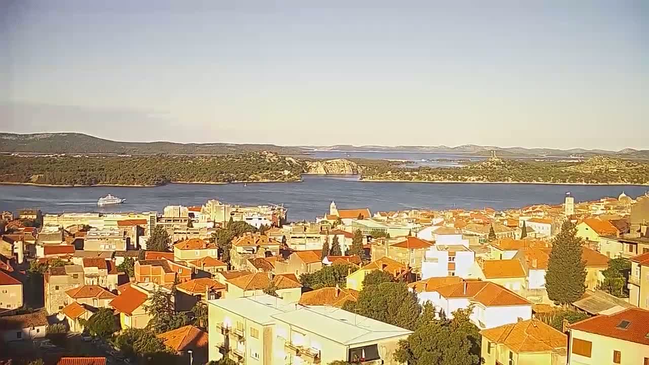 A coastal town with red-tiled roofs overlooks a calm bay surrounded by hills and islands under a bright, sunny sky.