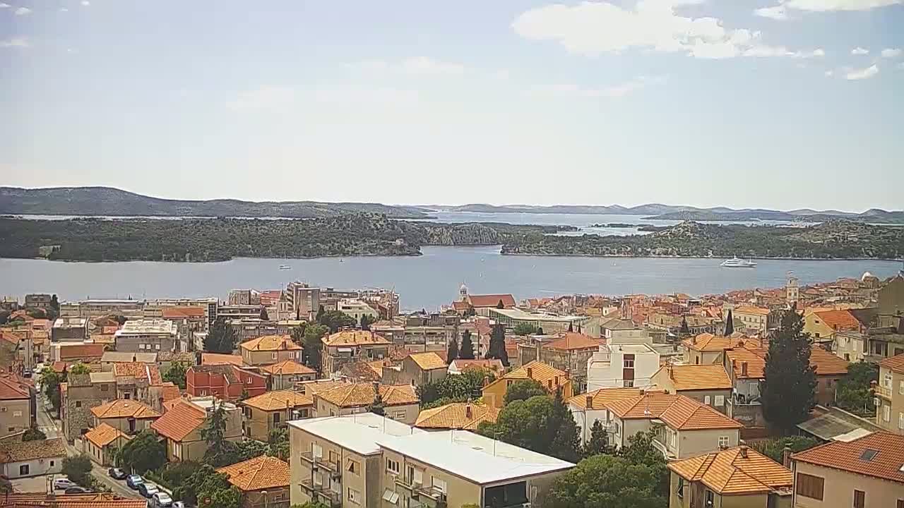 A coastal town with predominantly orange-roofed buildings stretches across a hillside overlooking a calm body of water under a partly cloudy sky.