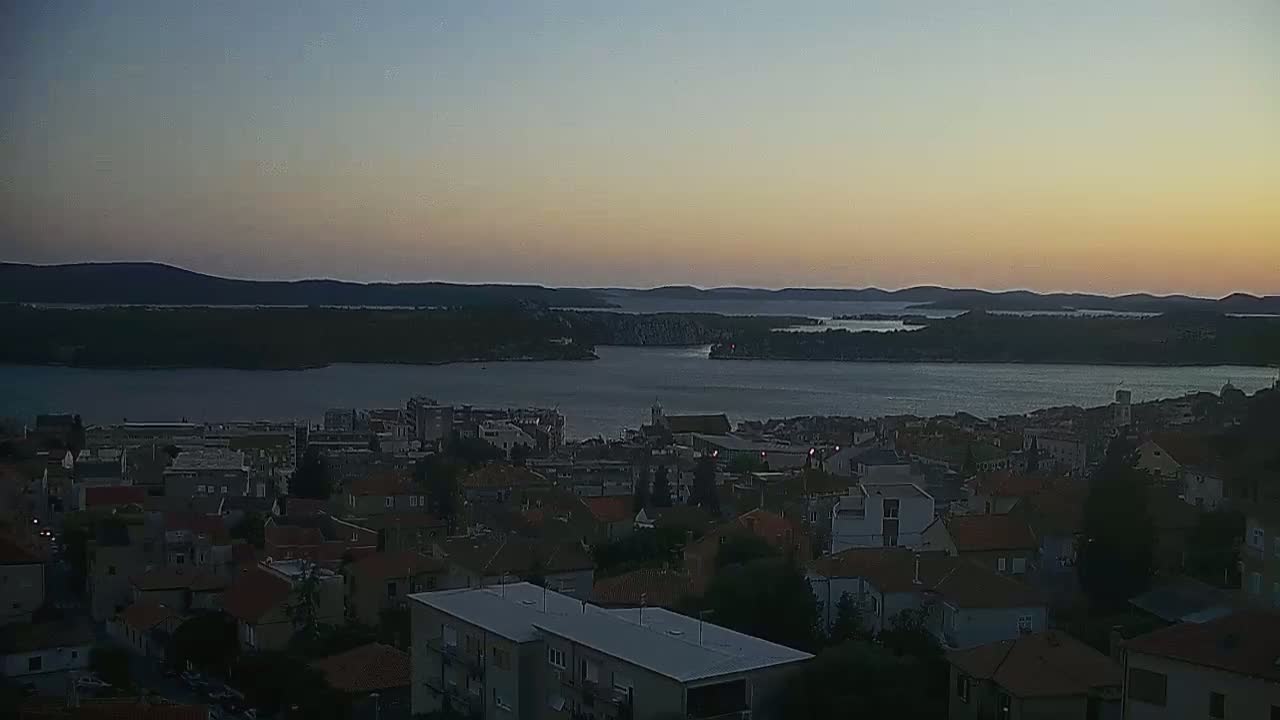 A coastal town with numerous houses and buildings overlooks a calm bay at sunset, with dark silhouettes of hills and islands in the distance under a clear sky.