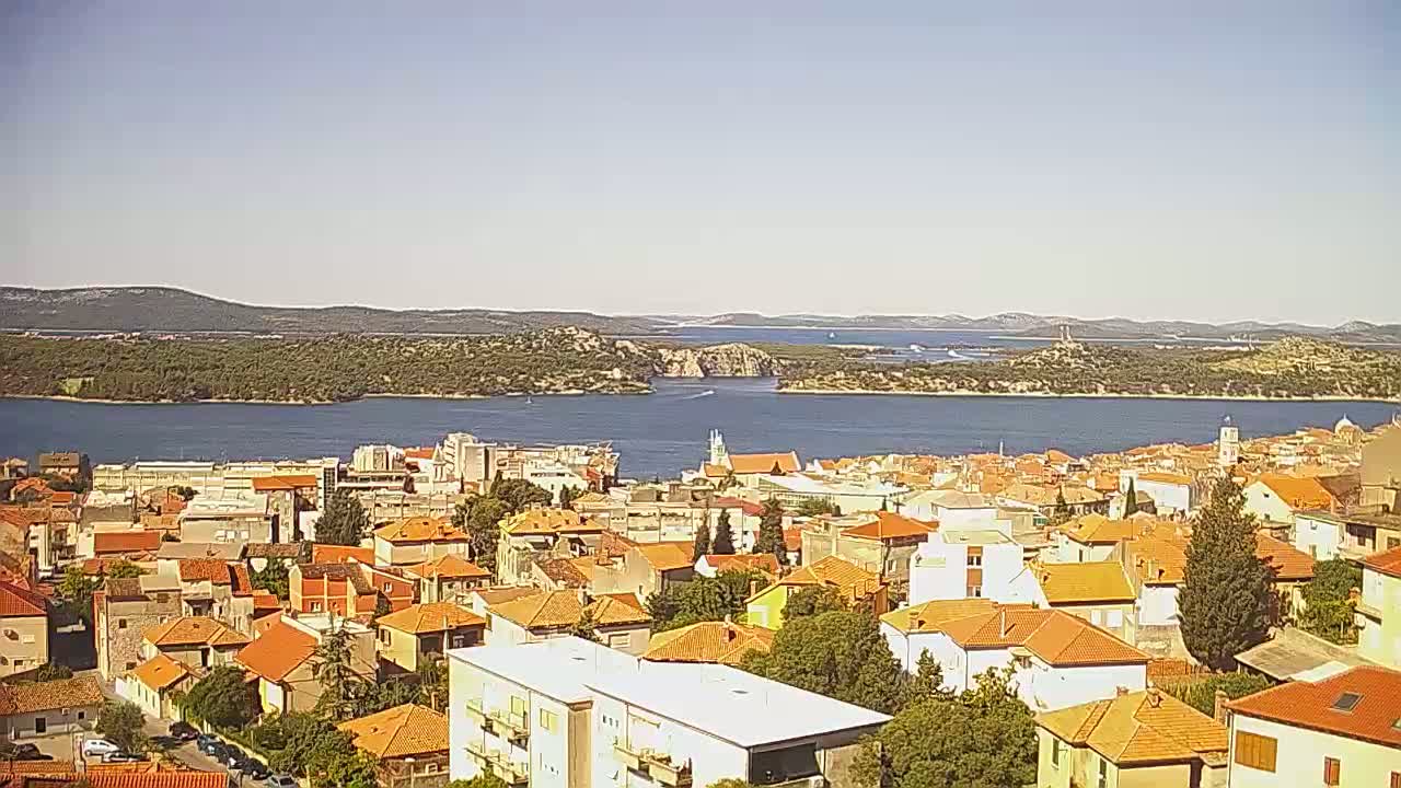A coastal town with mostly orange-roofed buildings stretches along a calm body of water under a clear, sunny sky, with distant islands visible across the bay.
