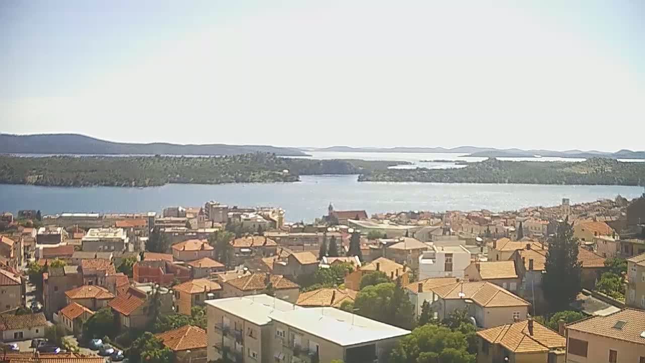 A coastal town with red-tiled roofs stretches across a hillside overlooking a calm bay dotted with islands under a clear, sunny sky.