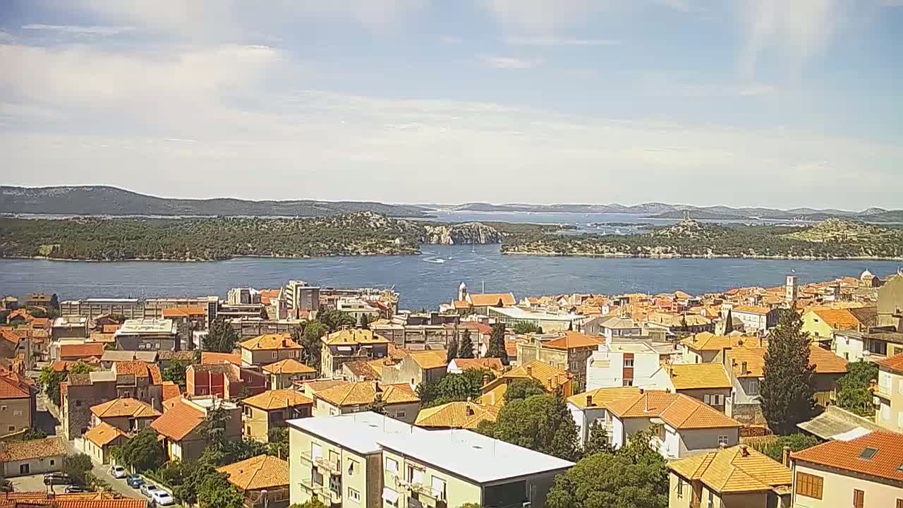 A coastal town with red-tiled roofs overlooks a calm body of water under a partly cloudy sky.
