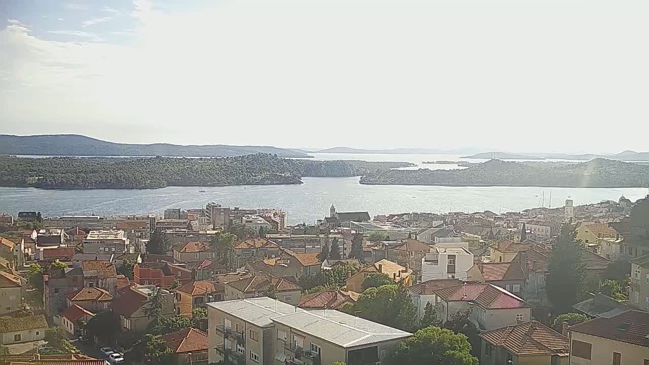A coastal town with red-tiled roofs sits on a hillside overlooking a calm bay and distant islands under a mostly sunny sky.