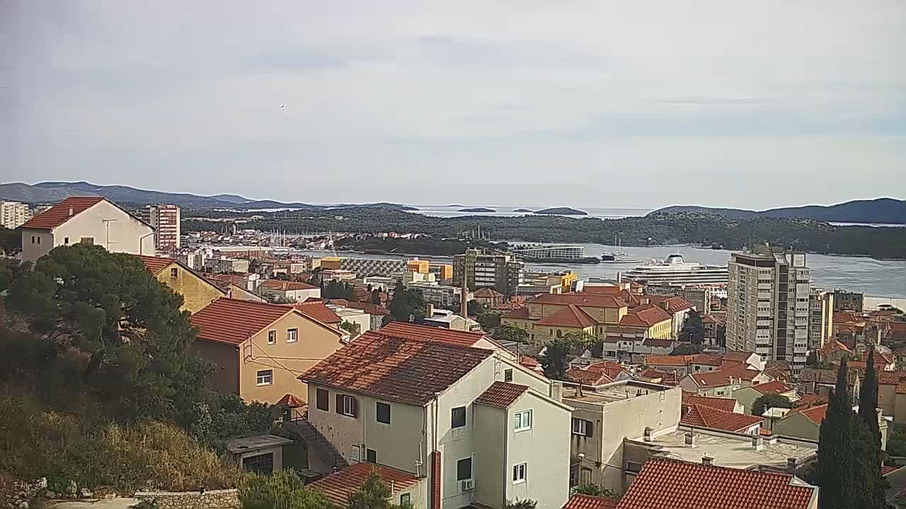 A coastal town with red-tiled rooftops, a large cruise ship in the harbor, and slightly overcast skies.