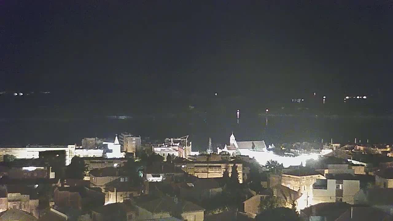 A nighttime view of a coastal town, illuminated by streetlights, with a calm body of water in the background under a dark sky.