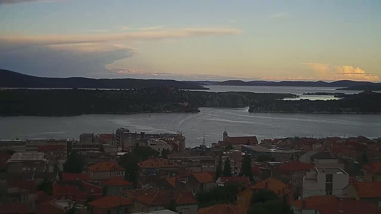 A coastal town with red-tiled roofs overlooks a calm bay under a partly cloudy, dusky sky.