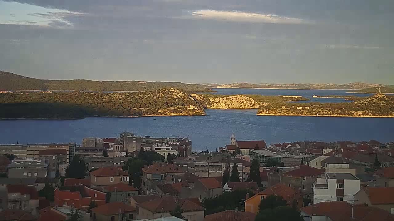 A coastal town with red-tiled roofs overlooks a calm bay and distant islands under a partly cloudy sky.