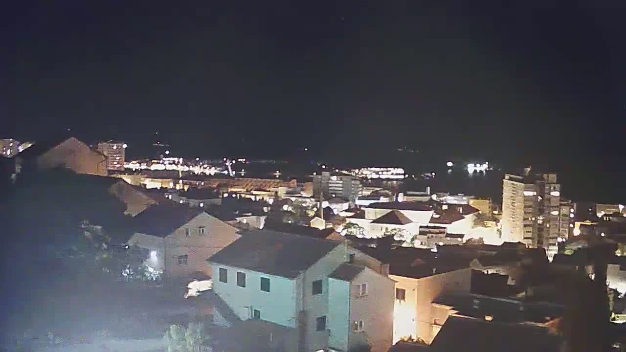 A nighttime, high-angle view of a town with numerous illuminated buildings, extending to a dark body of water in the distance under a clear, dark sky.