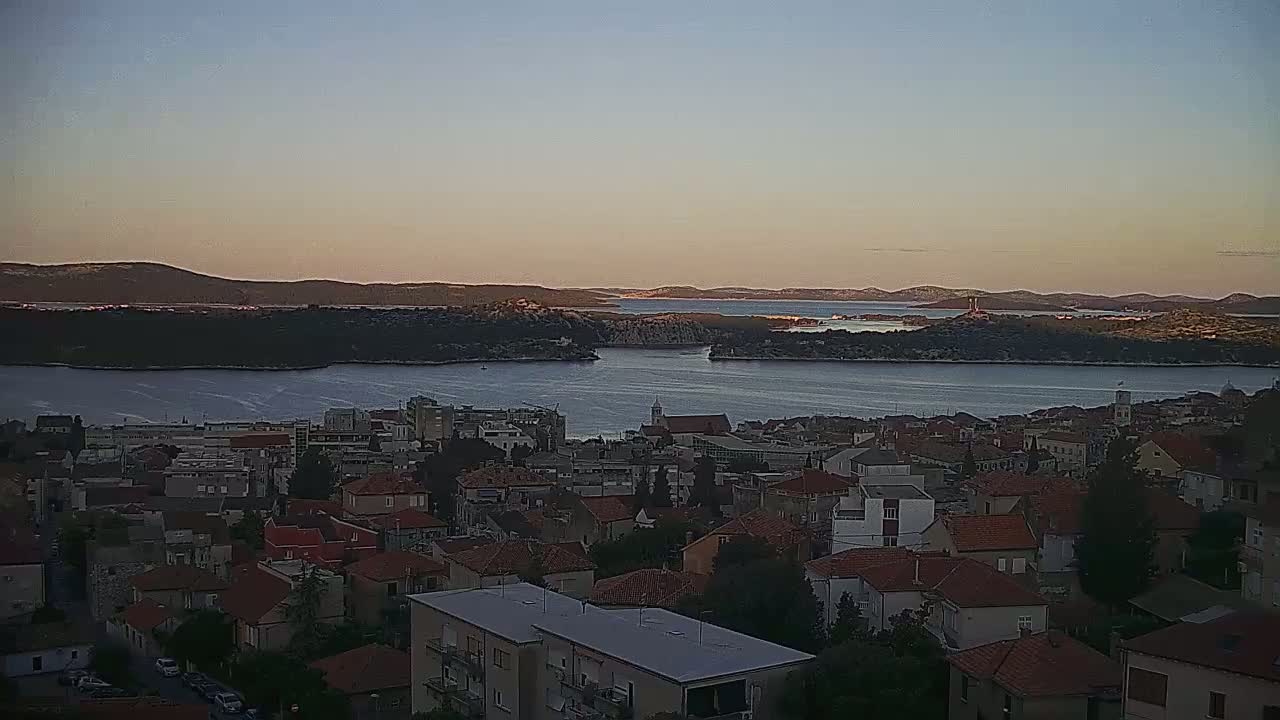 A coastal town with red-tiled roofs overlooks a calm body of water at sunset, with islands and hills visible in the distance.