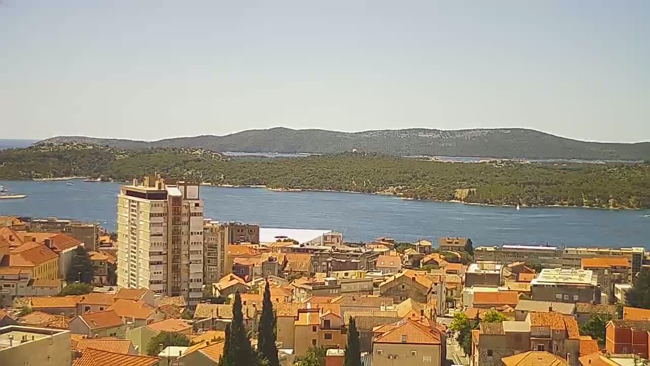 A coastal town with red-tiled roofs and a tall building overlooks a calm body of water and a green island under a clear sky.