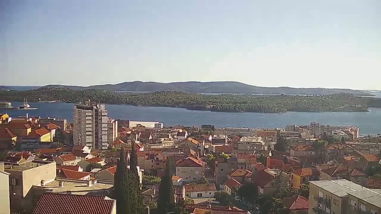 A coastal town with red-tiled roofs stretches towards a calm body of water under a clear, sunny sky, with a verdant island visible in the distance.