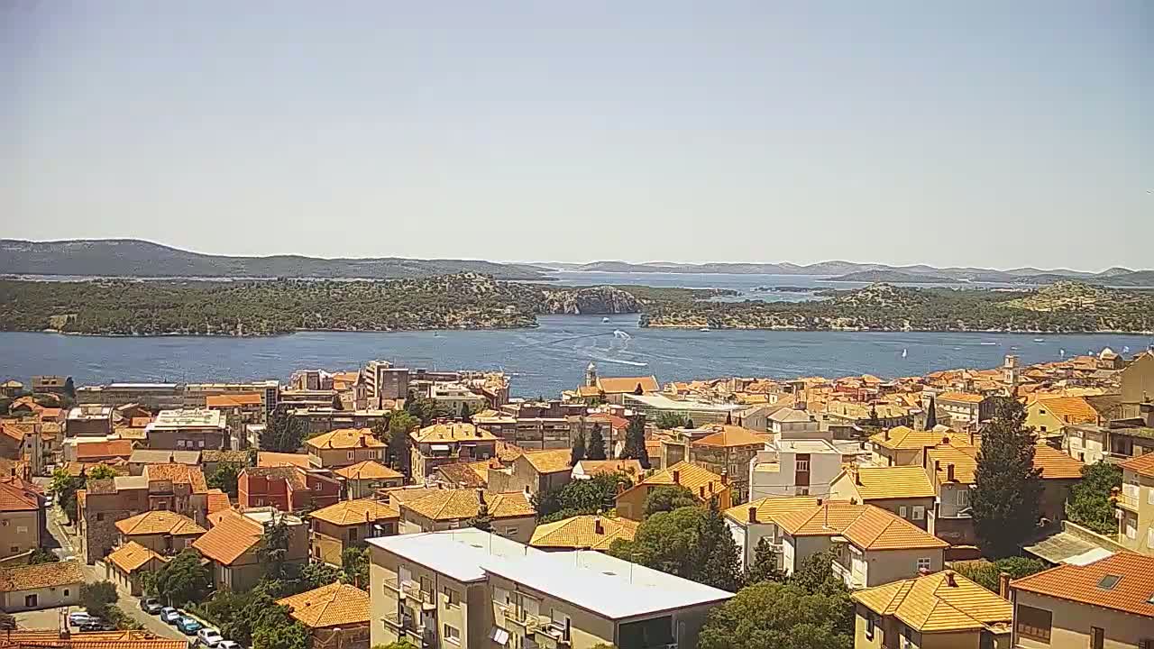 An elevated view shows a coastal town with numerous orange-roofed buildings, overlooking a calm body of water under a clear sky.