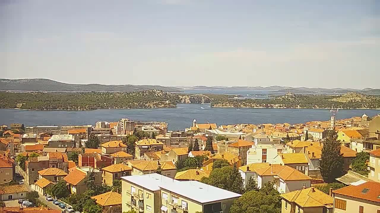 A coastal town with orange-roofed buildings stretches along a calm bay under a clear, sunny sky, with islands and hills visible in the distance.