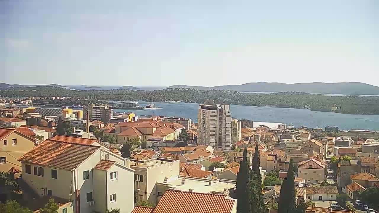 A coastal town with red-tiled roofs, a multi-story building, and a body of water under a clear, sunny sky.
