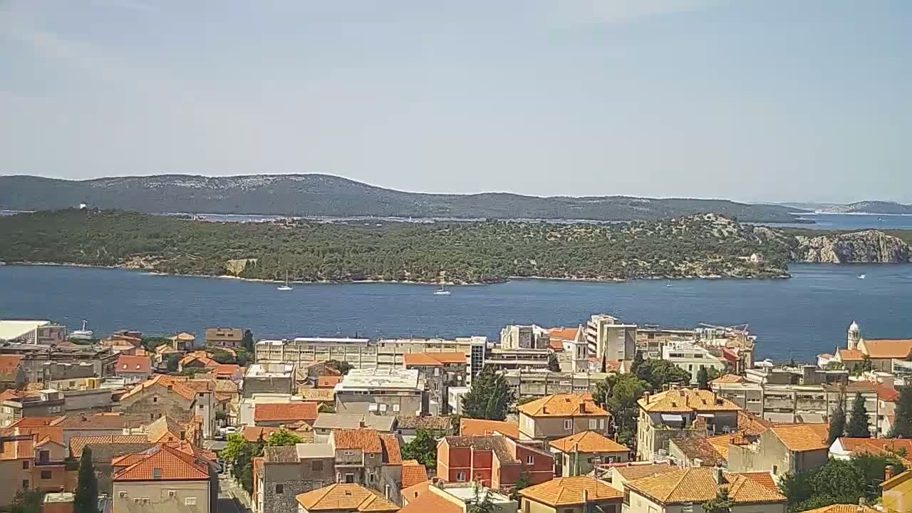 A coastal town with red-tiled roofs overlooks a calm bay and a verdant island under a clear sky.