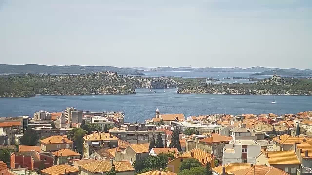 A town with many orange-roofed buildings overlooks a calm body of water and several islands under a clear sky.