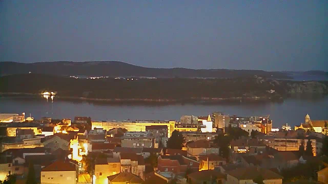 A nighttime view of a coastal town illuminated with warm lights, overlooking a calm body of water and a dark, tree-covered island in the distance under a twilight sky.
